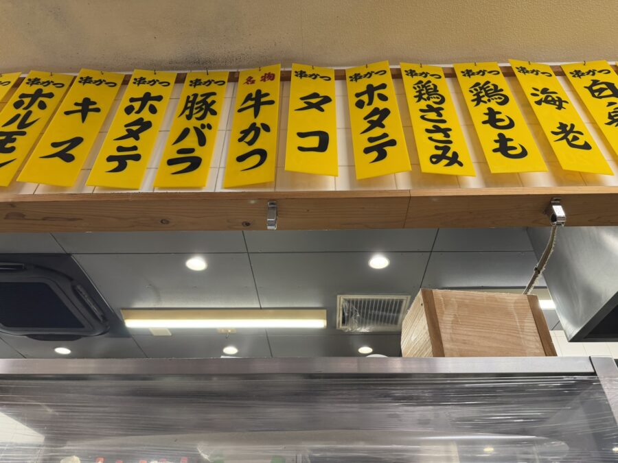 串カツ店に並ぶ黄色いメニュー札。ホルモン・キス・ホタテ・豚バラ・牛かつ・タコなど